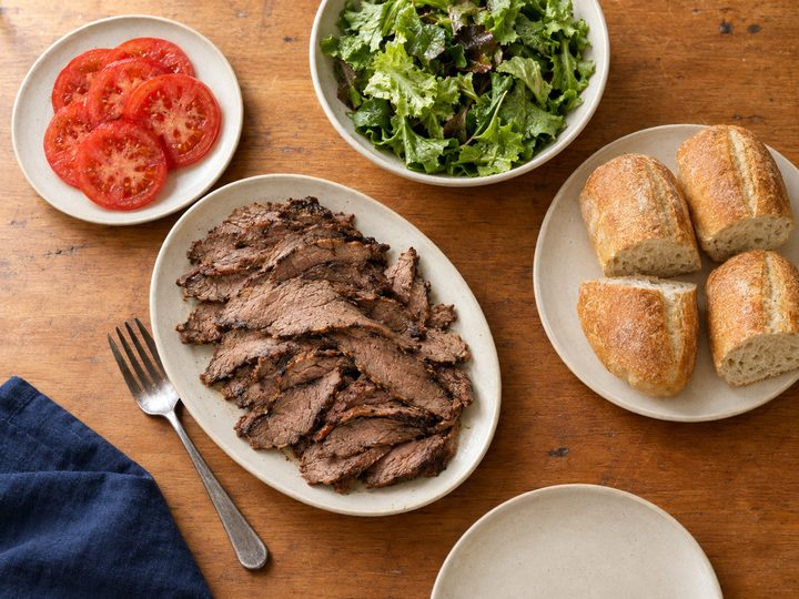 Overhead view of sliced cooked tri-tip, sliced tomatoes, a leafy green salad and crusty bread on separate cream ceramic dishes on a warm wooden table, with a navy napkin, serving fork and an empty plate at the edge.