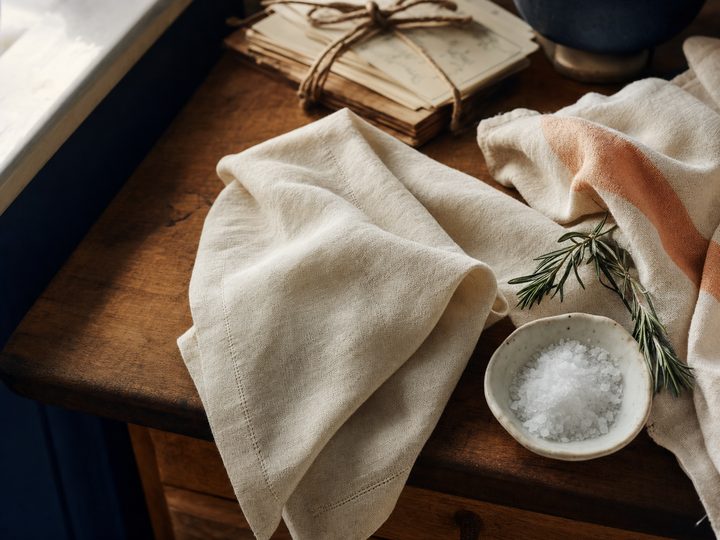 A cream linen towel with a peach stripe draped across a warm wooden counter beside a small dish of flaky sea salt, fresh rosemary, and a stack of handwritten recipe cards tied with twine.