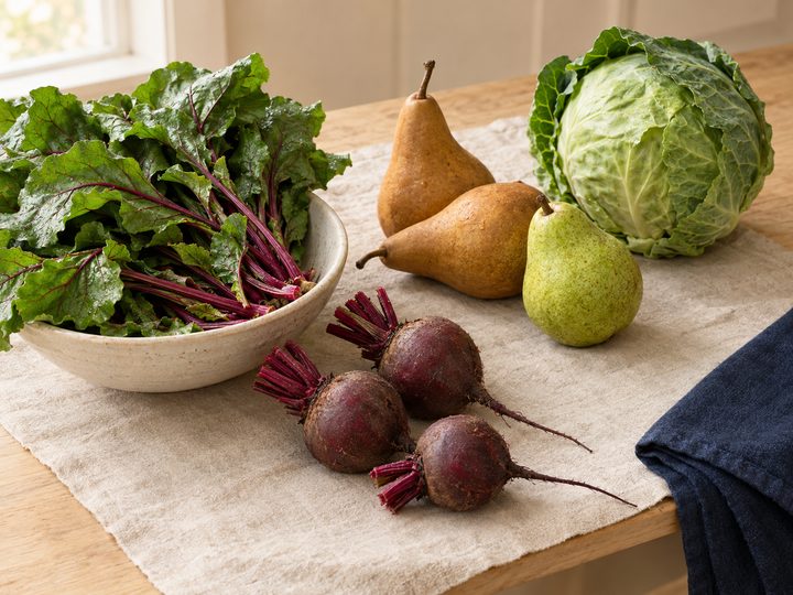 Beet greens in a cream ceramic bowl beside burgundy beet roots, russet and green pears, and a whole green cabbage on a pale wooden table with cream linen and a navy cloth.